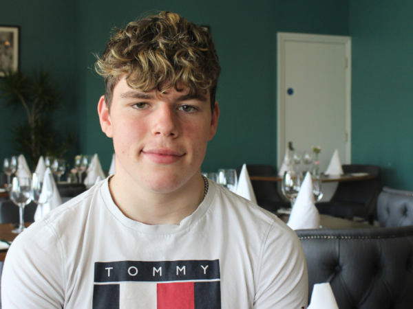 Student sitting on a chair in a restaurant. He is smiling at the camera and has mousey brown wavy hair, he is wearing a white t-shirt.
