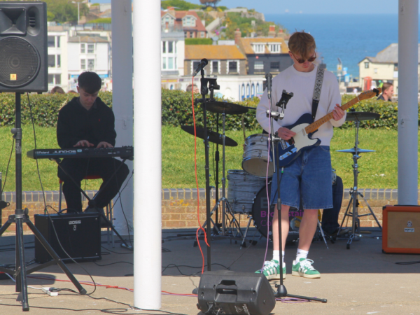 Two students performing on the Broadstairs bandstand. One student is playing a guitar wearing blue shorts and a white top, the other is dressed in black playing the keyboard.