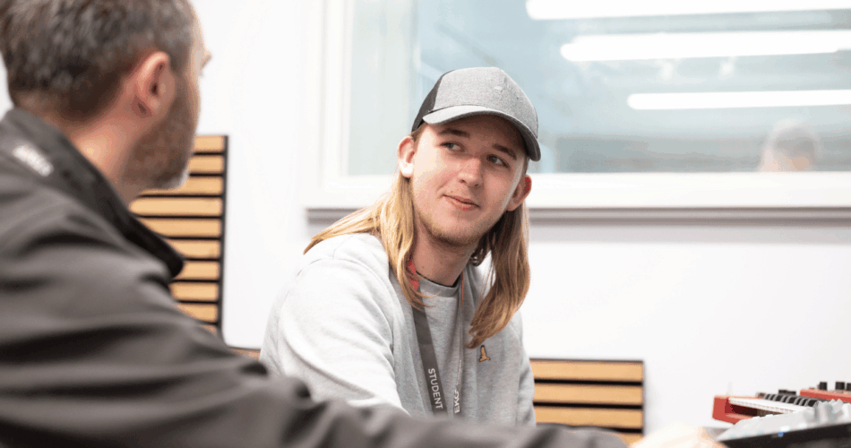 student with long blonde hair wearing a basketball hat looking at tutor next to him in a studio room.
