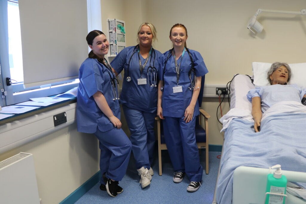 3 students in scrubs by a hospital bed with a life-sized doll 'patient'