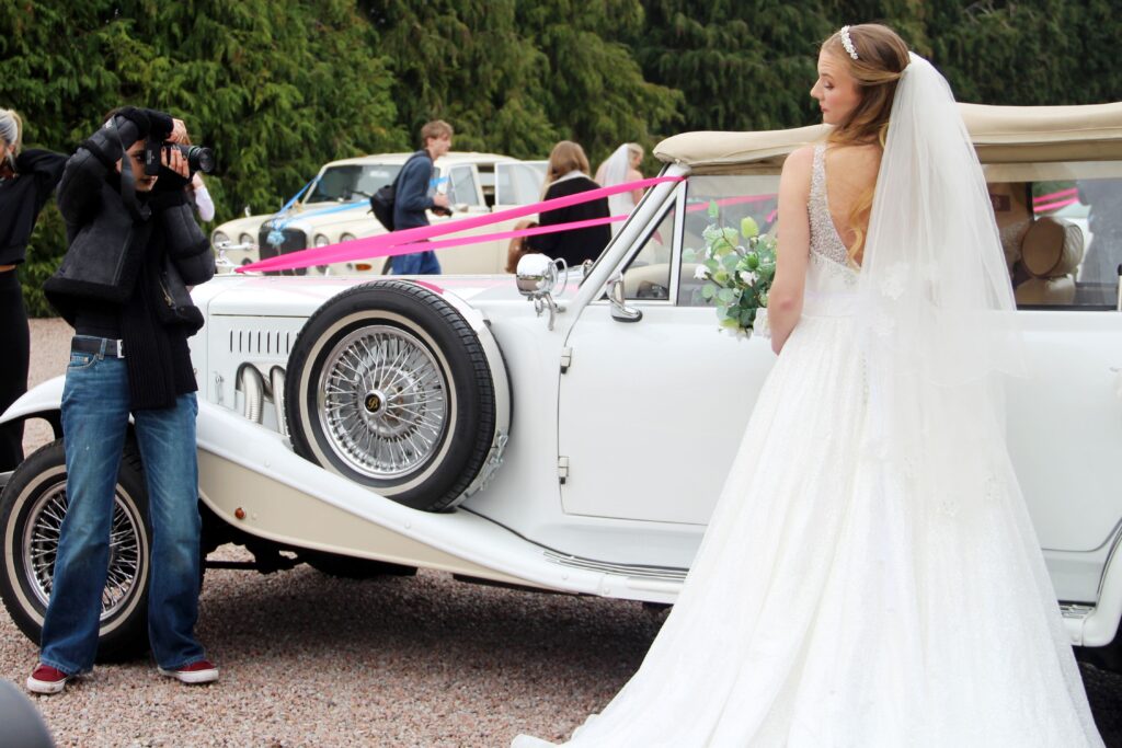 student photographer taking a photo of a model bride next to a vintage white car