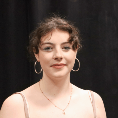 Student is smiling at the camera, she has brunette hair that is in a ponytail. Wearing gold hoop earrings and a gold necklace.
