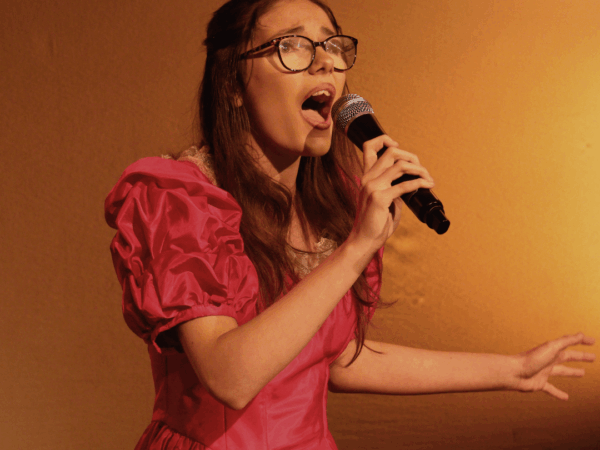 Student singing into microphone, she is wearing glasses and has brunette hair. She is wearing a red dress and the lighting is also red/orange.