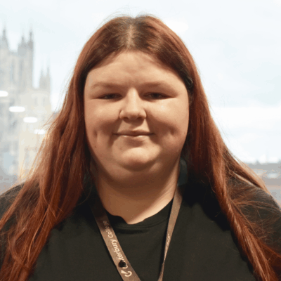 Student has long red hair and is wearing a black hoodie. She is smiling at the camera with a background of Canterbury Cathedral.