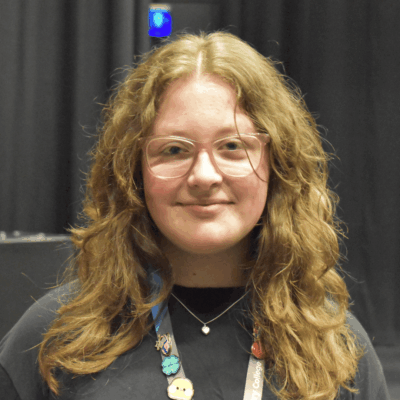 Student smiling at the camera, she has mousey brown shoulder length hair, glasses and is wearing a dark grey top.