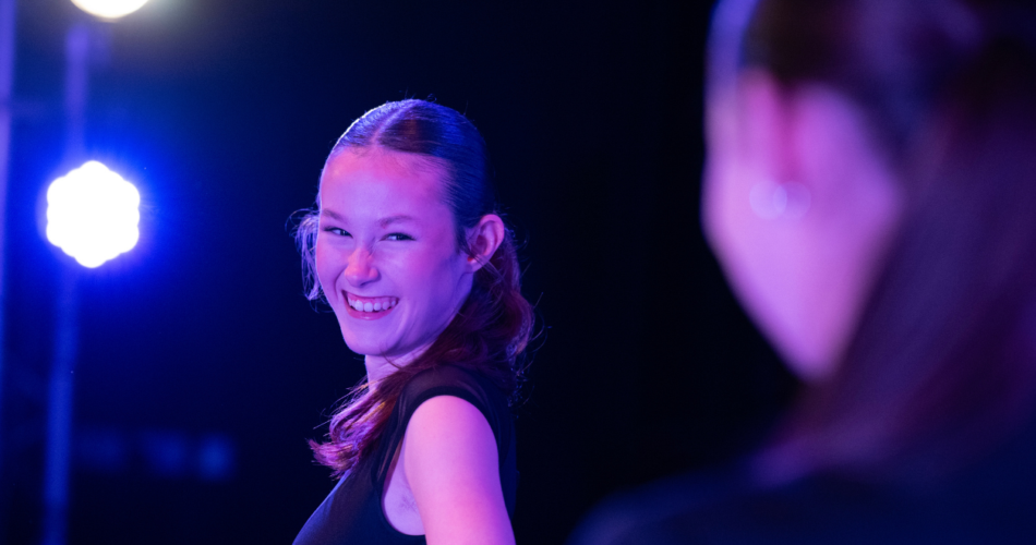 Student on a dark stage, close up of her looking over her shoulder laughing, she has her brunette hair in a ponytail and is wearing black.
