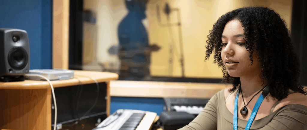 Student sitting at soundboard desk. She has black curly hair and is wearing a tan colours top.