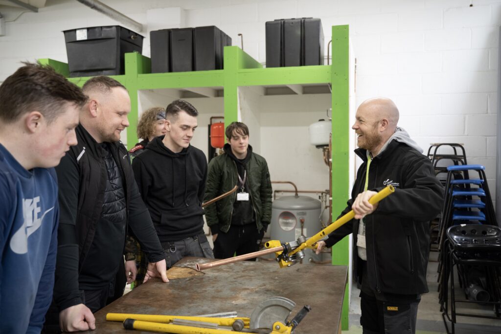Students watching a lecturer talking to them over a workbench holding a pipe.