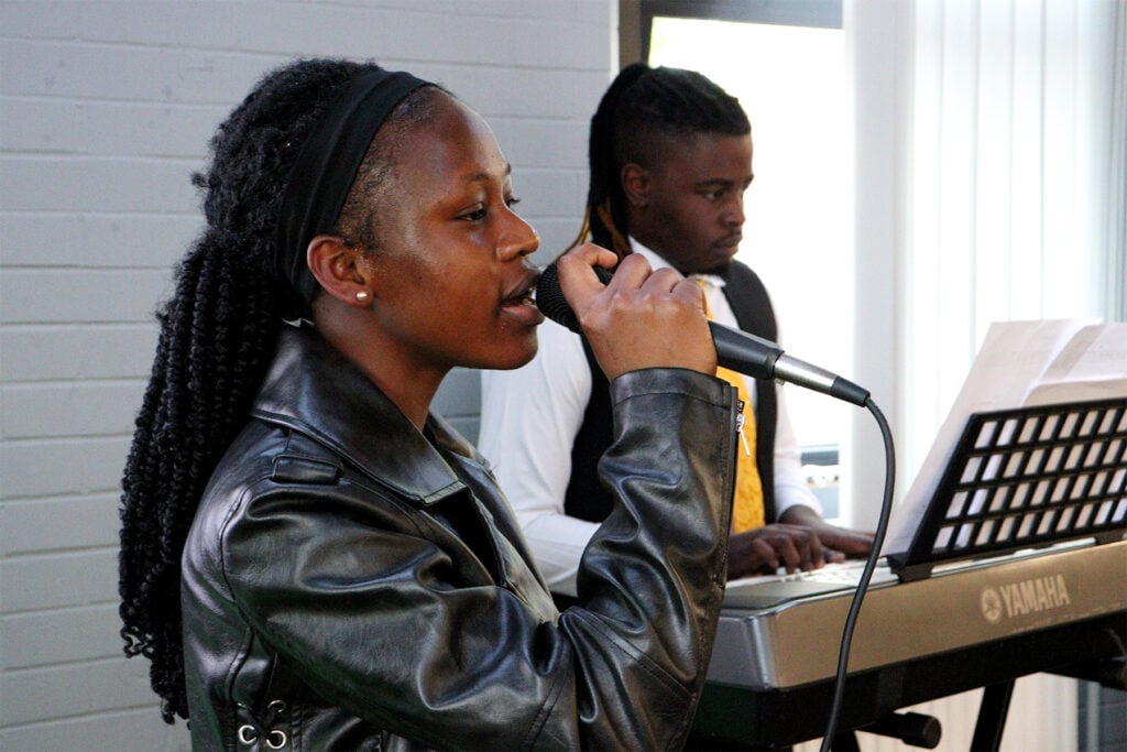 A student singing into a microphone with another student behind playing the keyboard