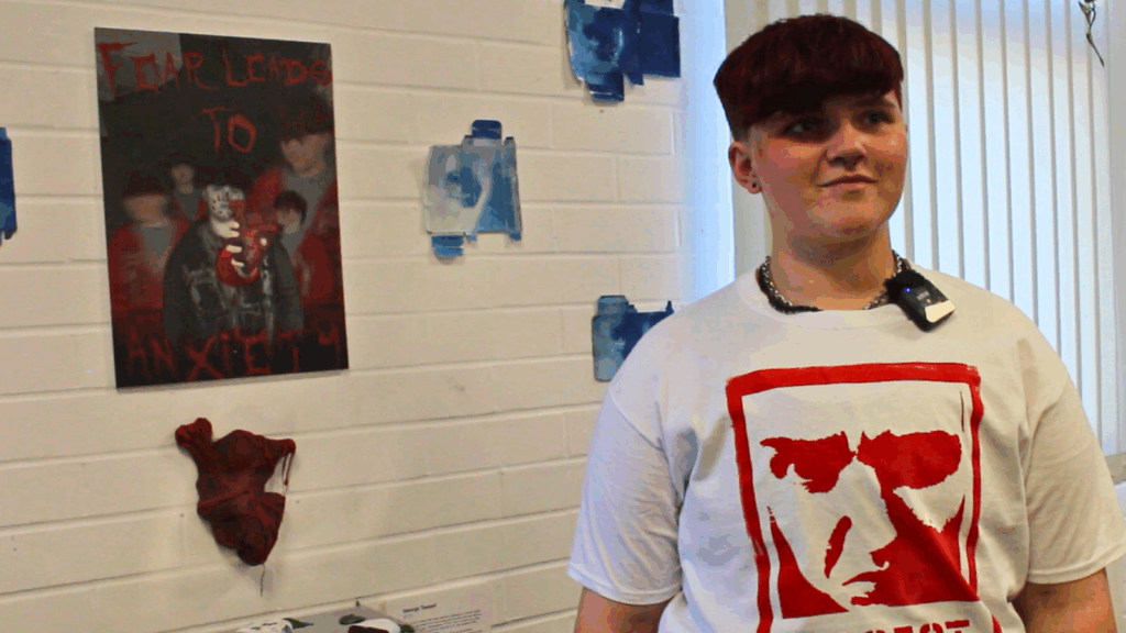 A photo of George and his artwork in the background of a canvas, pillboxes that have been coloured and have eyes on them and a red heart stuck to the wall.