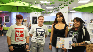 4 students looking at the camera smiling holding the Cene magazine showing their article within. Behind them is the Dover Art Exhibition