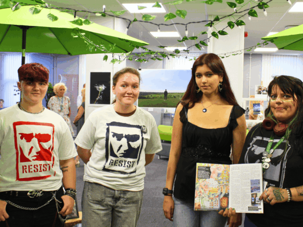 4 students looking at the camera smiling holding the Cene magazine showing their article within. Behind them is the Dover Art Exhibition