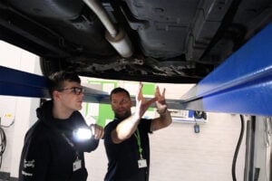 2 people under a car which is on a ramp. one person is holding a torch.