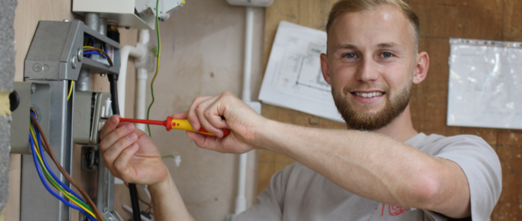 student smiling at camera whilst holding a screwdriver and working on electrical plug,