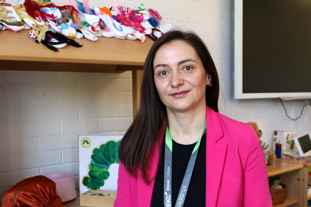 Adult learner with long black straight hair wearing a black t-shirt and pink blazer looking and smiling at the camera. There is a children's bookcase behind her.