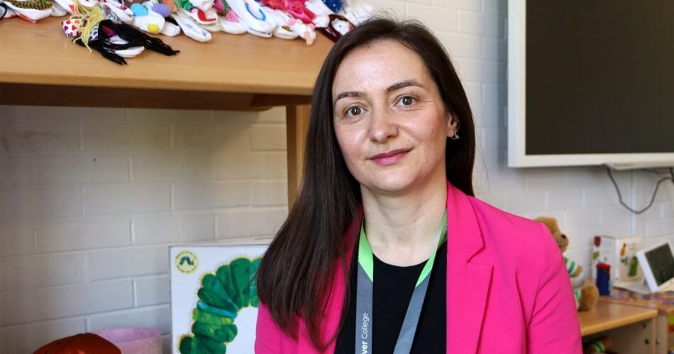 Adult learner with long black straight hair wearing a black t-shirt and pink blazer looking and smiling at the camera. There is a children's bookcase behind her.