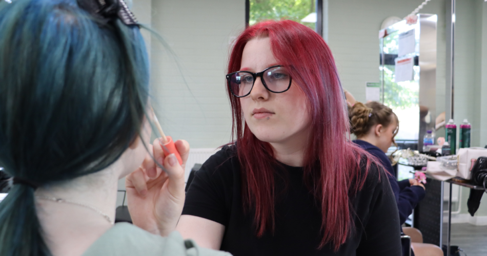 Student has long red hair and wearing black glasses, she is putting concealer onto another woman's face.