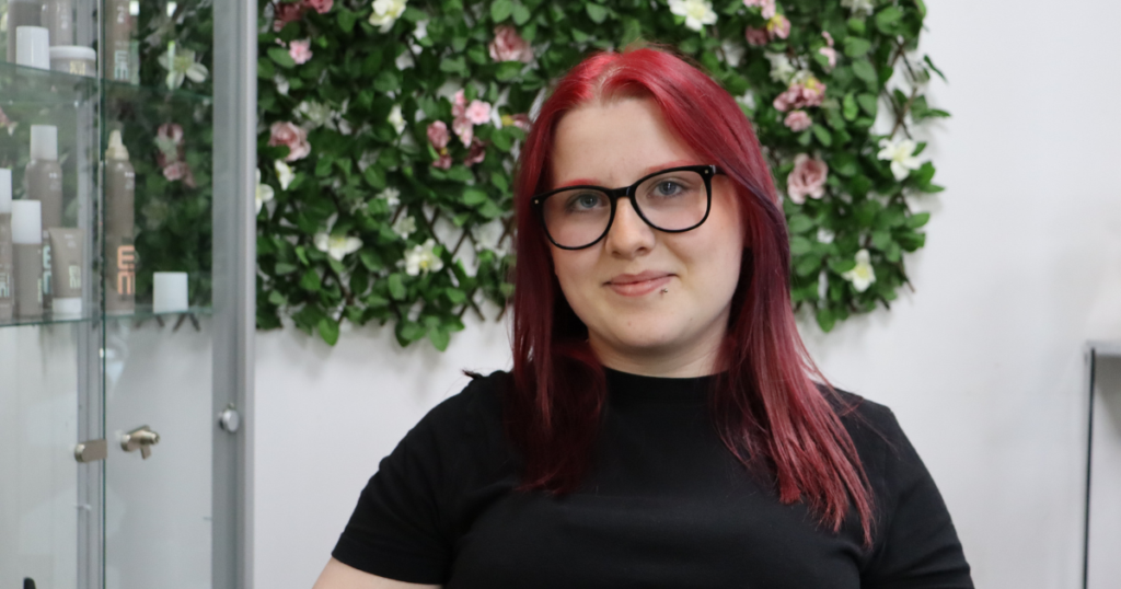 Student smiling at camera sitting on a chair with a flowery background. She has dark red hair and wears black glasses, she also has a black top on.