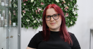 Student smiling at camera sitting on a chair with a flowery background. She has dark red hair and wears black glasses, she also has a black top on.