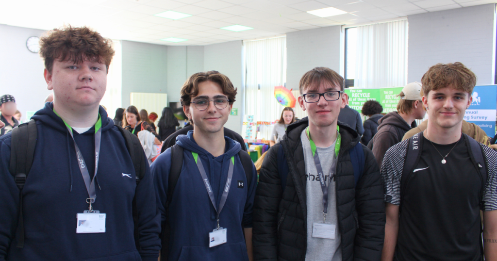 Four male students standing in front of the freshers fayre smiling at the camera