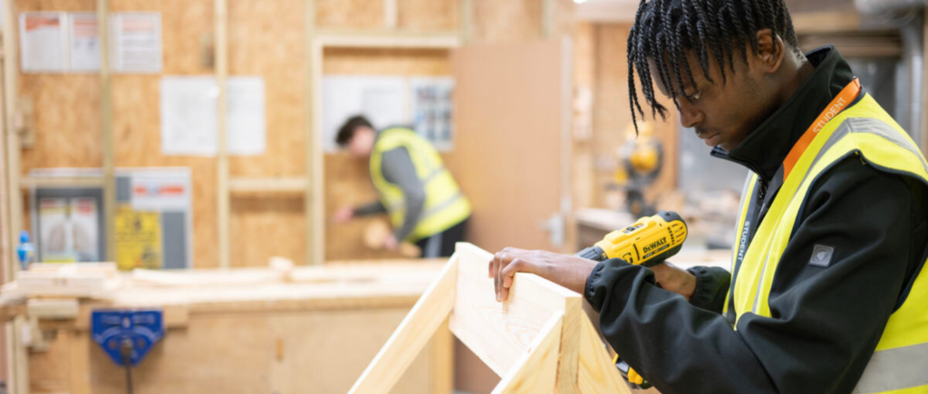 Student using drill to create a hole in a wooden frame structure.