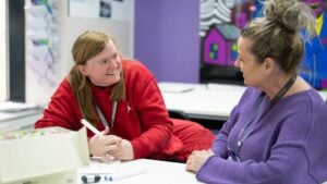 Student smiling and talking to a teacher at a table in a classroom.
