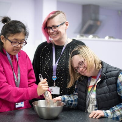 Two inclusive learners mixing a bowl of ingredients in a kitchen with a lecturer, smiling.