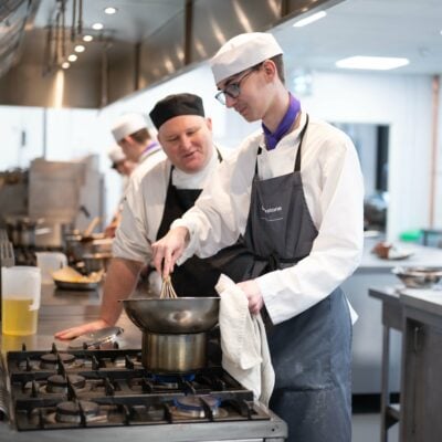 student cooking over hob with lecturer helping. Wearing aprons and smiling.