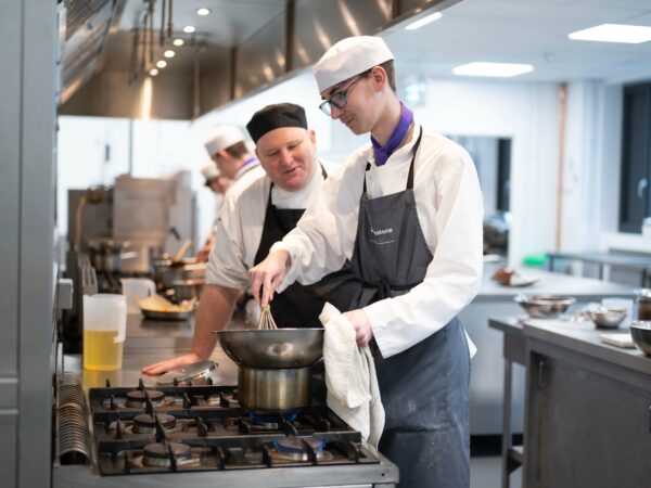 student cooking over hob with lecturer helping. Wearing aprons and smiling.