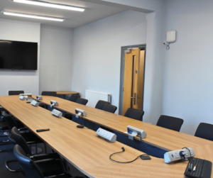 Photo of a meeting room with chairs and a long table. At the end is a big tv on the wall.