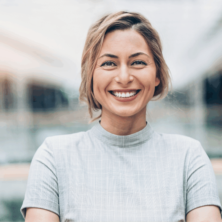 Woman smiling at the camera. She is wearing a light grey top and is standing outside.