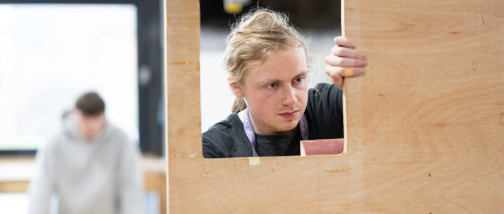Student looking through square cut-out in a piece of wood with a tool.