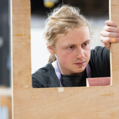 Student looking through square cut-out in a piece of wood with a tool.