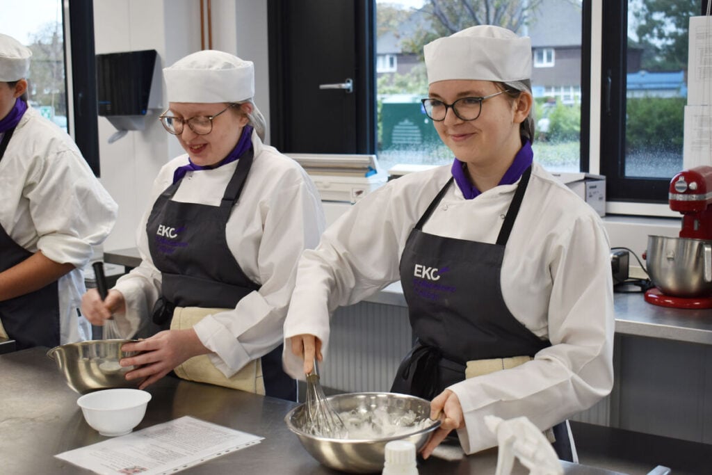 Two students with catering hat and apron on whisking eggs in a metal bowl, smiling.