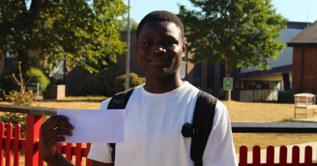 Student smiling holding grades outside. He is wearing a white top.