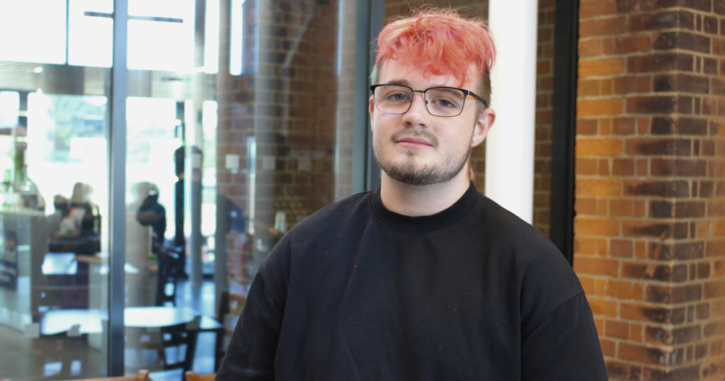 Student is wearing glasses, a black top and has pinkish hair. He is smiling at the camera.