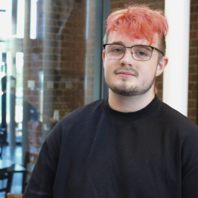 Student is wearing glasses, a black top and has pinkish hair. He is smiling at the camera.