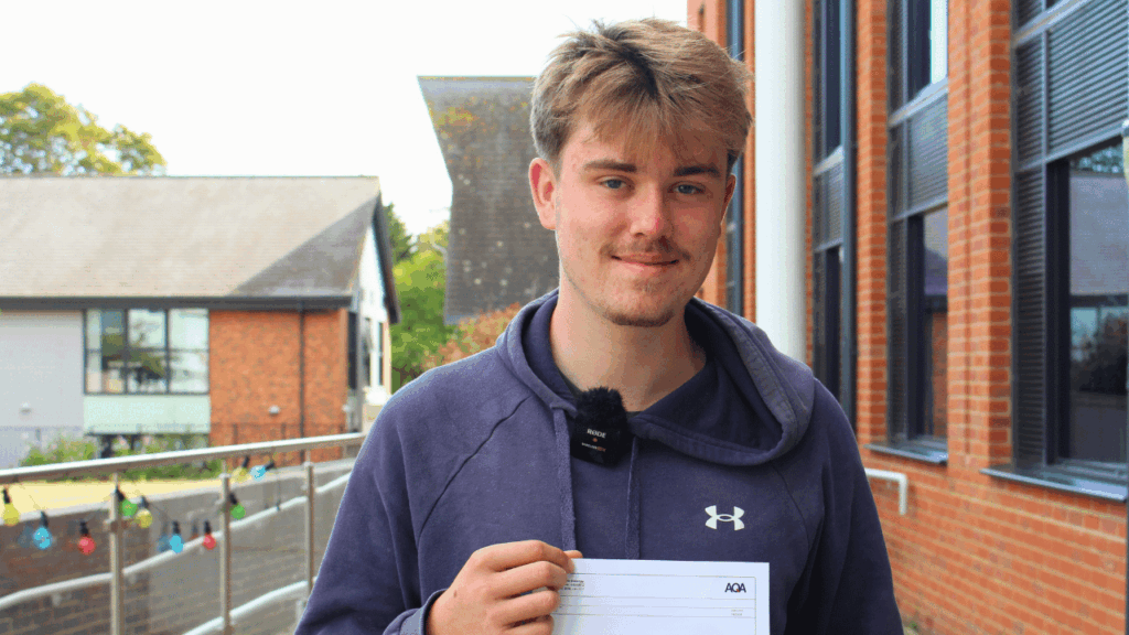 Student holding up grade paper, he is wearing a blue hoodie and has mousey brown hair.