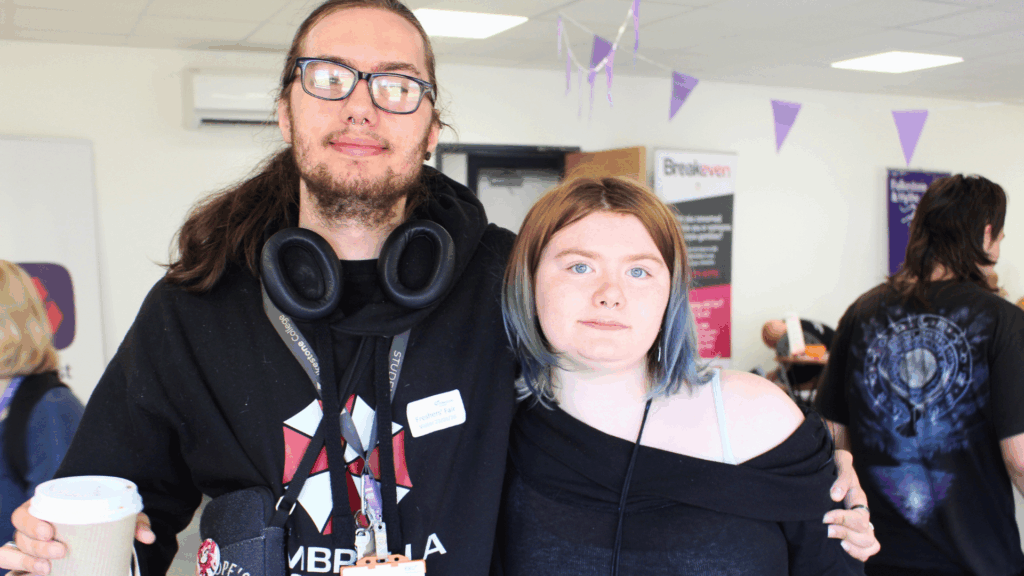 Student wearing glasses and a black hoodie holding a coffee and another student with shoulder length hair and blue eyes wearing a black top. They are both smiling at the camera.