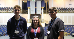 Three business students standing on the stage in the grand hall of Leas Cliff Hall. They are smiling at the camera.