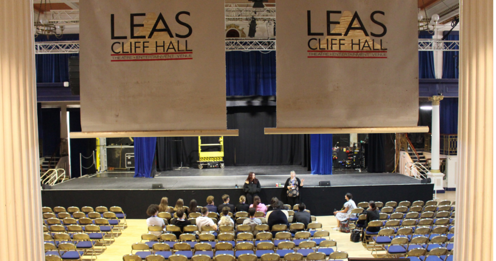 Inside the grand hall of Leas Cliff Hall looking at the seated stalls area and the stage. The business students are seated in the middle. There are Leas Cliff Hall banners hanging down and pillars either side of the photo